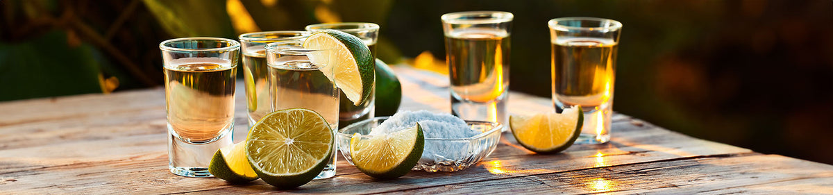 Sechs Tequila-Shots mit Limettenspalten und einer Schale Salz auf einer hölzernen Unterlage in warmem Tageslicht.