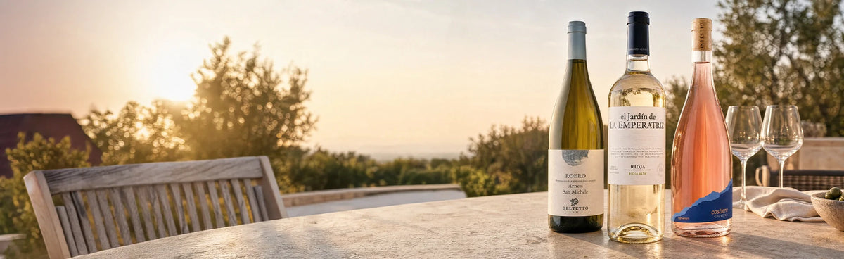 Drei Flaschen Wein und zwei leere Gläser auf einem Tisch im Freien bei Sonnenuntergang, mit Bäumen und Stühlen im Hintergrund.