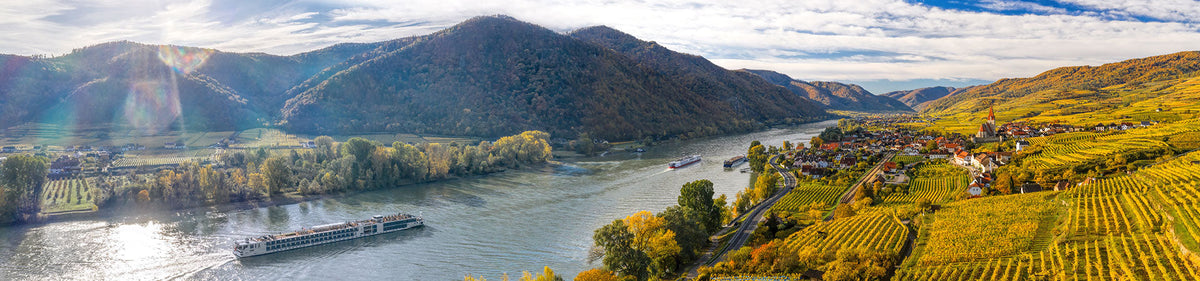 Panoramablick auf ein Flusskreuzfahrtschiff, das unter blauem Himmel an Weinbergen und einem kleinen Dorf inmitten grüner Hügel vorbeifährt.