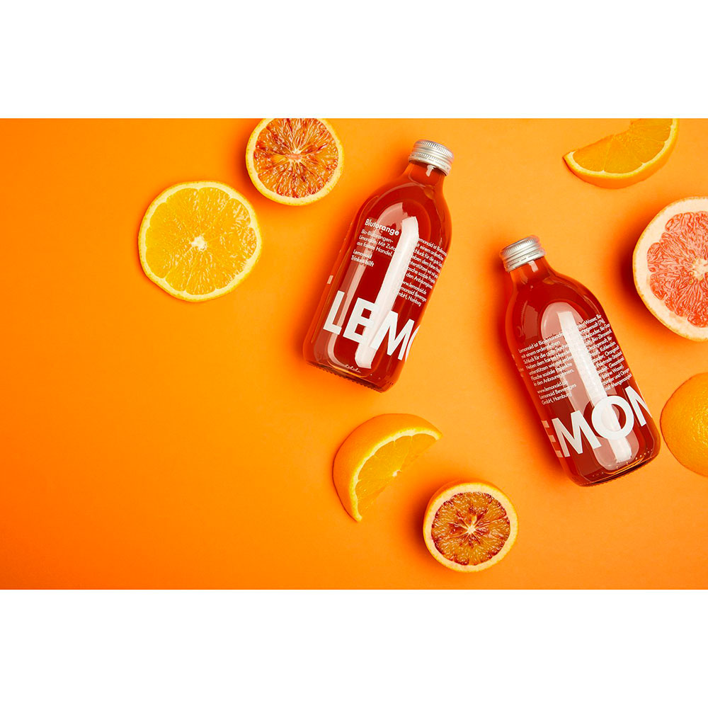 Zwei Flaschen Lemonaid Blutorange 0,33L auf einem orangefarbenen Hintergrund mit Zitrusscheiben, von oben betrachtet - ein lebhaftes Schaufenster für die fair gehandelten Bio-Getränke von Lemonaid.