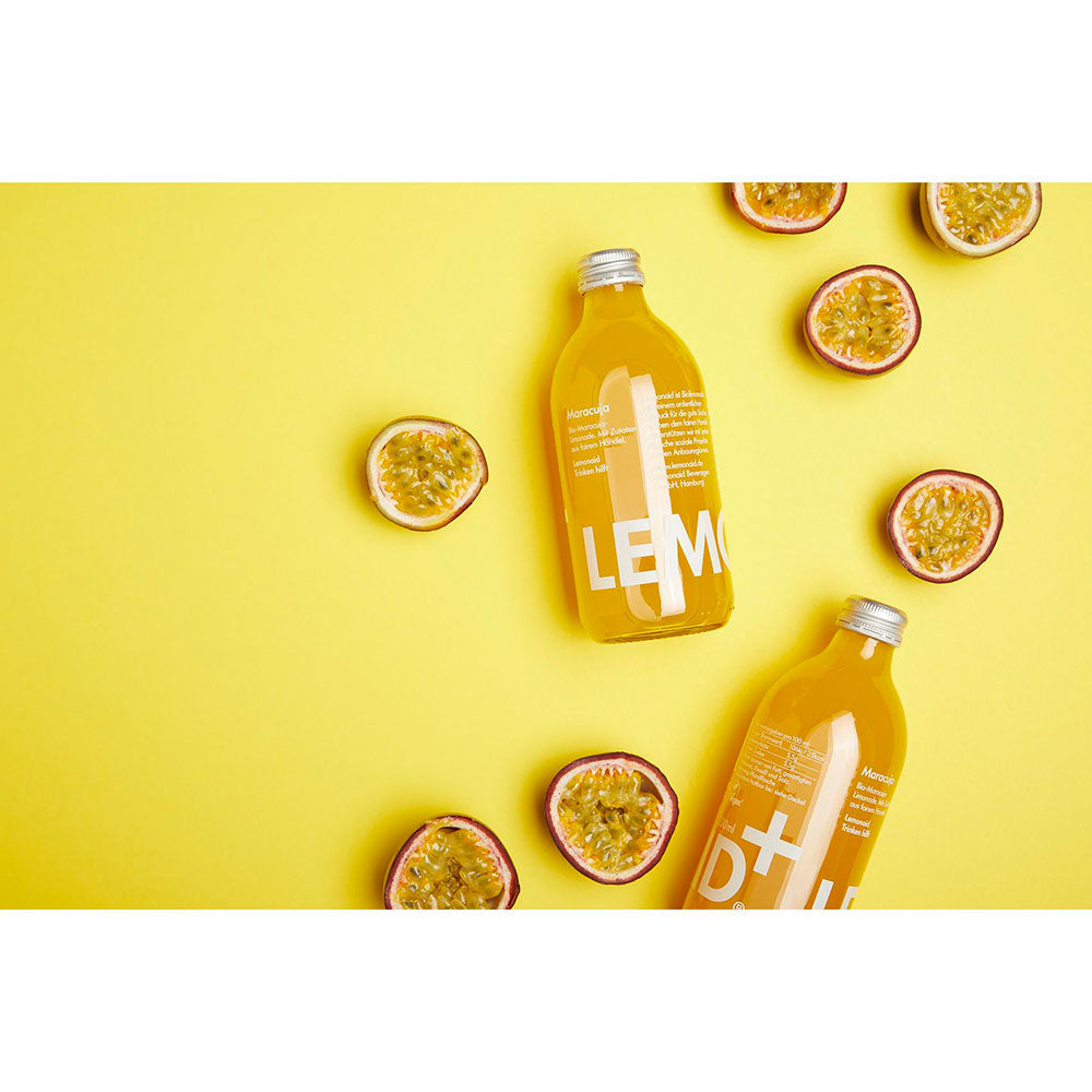 Zwei Flaschen Lemonaid Maracuja 20x 0,33 Liter (MEHRWEG) von Lemonaid und halbierte Passionsfrüchte sind auf einem leuchtend gelben Hintergrund zu sehen.