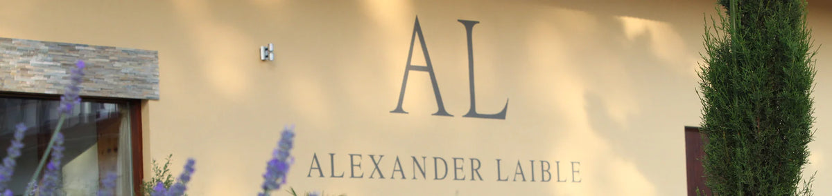 Außenwand eines Gebäudes mit der Aufschrift AL Alexander Laible, teilweise umrahmt von Pflanzen.