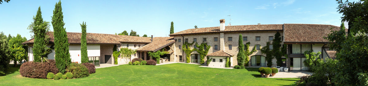 Eine große, rustikale Villa mit Steinmauern, Bogenfenstern und Ziegeldächern, umgeben von Bäumen und grünen Rasenflächen.