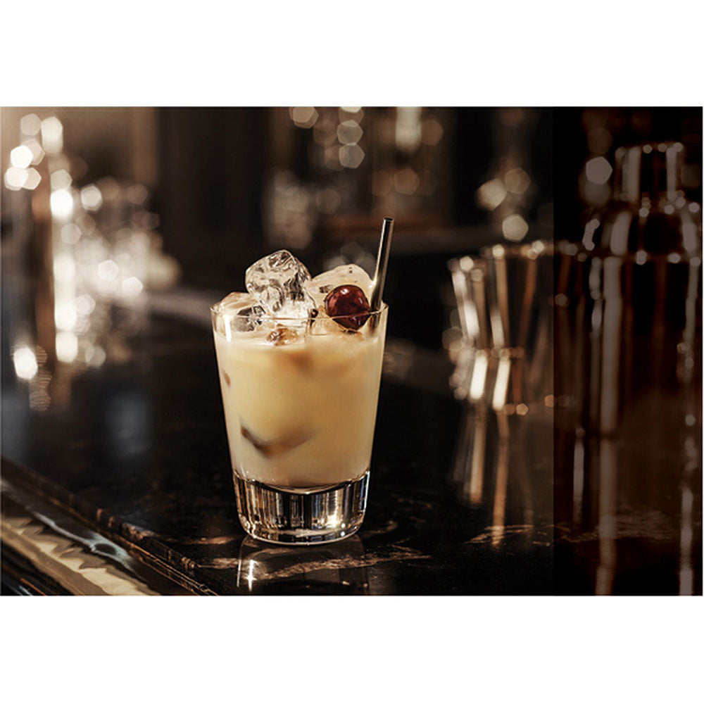 Ein White Russian mit Eis, Kirsche und Strohhalm - angereichert mit einem Spritzer Baileys Irish Cream 17% 0,7L von Baileys - steht auf einem dunklen Bartresen vor einem verschwommenen Hintergrund.