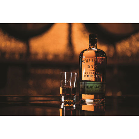Eine Flasche Bulleit 95 Rye Frontier Whiskey 45% (0,7 l) und ein Whiskeyglas ruhen auf einer reflektierenden Oberfläche unter warmer Beleuchtung, die die Qualität von Bulleit Frontier Whiskey hervorhebt.